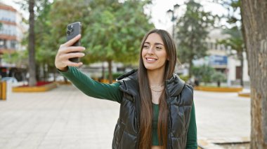 Gülümseyen bir kadın şehir parkında ağaçlar ve banklarla selfie çeker, şehir dışı yaşam tarzını gözler önüne serer..