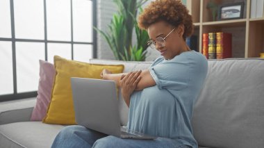 Rahat bir Afrikalı Amerikalı kadın modern bir oturma odasında dizüstü bilgisayarını kullanırken kolunu uzatıyor..