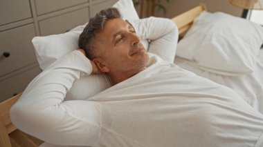 Orta yaşlı bir adam evde yatakta dinleniyor. İyi aydınlatılmış bir yatak odasında mutlu ve rahat görünüyor..
