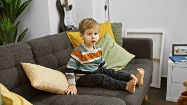 Modern bir oturma odasında gri bir koltukta çıplak ayakla oturan beyaz çocuk, düşünceli görünüyor..