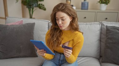 Oturma odasındaki güzel sarışın bir kadın elinde kredi kartı tutarken mavi bir tablet kullanıyor. İnternet üzerinden alışveriş yaptığını ya da internette gezindiğini gösteriyor..