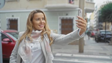 Gülümseyen sarışın kadın şehrin hareketli caddesinde selfie çekiyor, kentsel yaşam tarzı ve teknolojisini somutlaştırıyor..