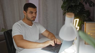 Evde dizüstü bilgisayar kullanan genç bir adam, iyi aydınlatılmış bir dairede beyaz bir masada otururken odaklanmış bir yüz ifadesi sergiliyor..