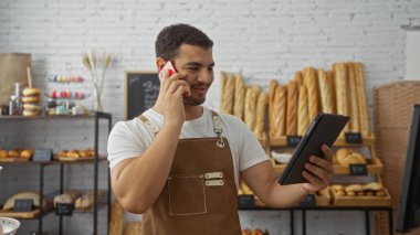 Fırındaki genç adam elinde tabletle telefonla konuşuyor. Raflar ekmek ve hamur işleriyle dolu.