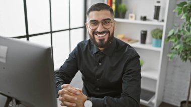 Sakallı genç bir adam modern ofis ortamında kameraya gülümsüyor, kendine güveni ve ulaşılabilirliği var..