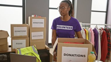 Afrikalı Amerikalı bir kadın, kapalı bir bağış merkezinde gönüllü olarak kutuları ve kıyafetleri düzenliyor..