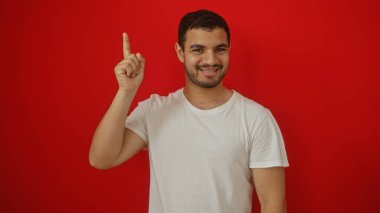 Genç bir adam canlı kırmızı bir arka plan karşısında beyaz bir gömlek içinde gülümsüyor, parmağıyla yukarıya doğru işaret ederek, parlak, izole bir ortamda güven ve pozitifliği resmediyor.