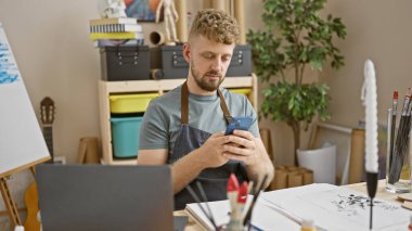 Mavi gözlü genç sakallı bir adam, sanat malzemeleriyle çevrili yaratıcı bir stüdyo ortamında akıllı bir telefon kullanır..