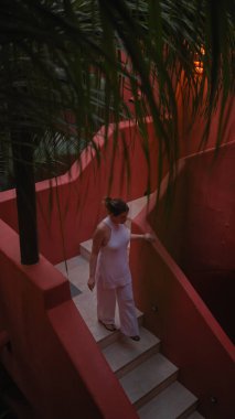 A young woman in white attire descends a vibrant red staircase surrounded by lush greenery at a tropical balinese resort. clipart