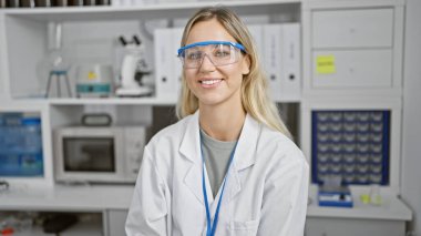 Laboratuvar gözlüğü ve beyaz önlük takan gülümseyen beyaz bir kadın laboratuvar ortamında güvenle duruyor..