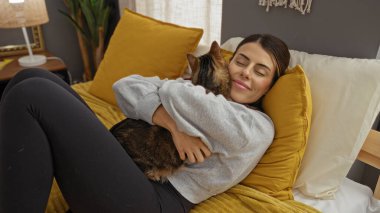 Genç bir İspanyol kadın evdeki rahat bir yatak odasında evcil kedi yavrusunu yatakta kucaklıyor..