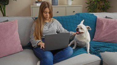 Genç sarışın bir kadın, rahat bir oturma odasında beyaz köpeğiyle kanepede otururken dizüstü bilgisayarda çalışıyor..