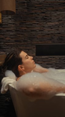 Huzurlu genç bir kadın modern bir banyo ortamında rahatlatıcı köpük banyosunun keyfini çıkarıyor..