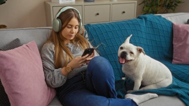 Genç sarışın bir kadın, rahat bir oturma odasında köpeğiyle kanepede dinlenirken kulaklıkla müzik dinliyor..