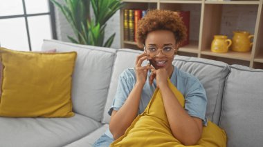 Çekici bir Afrikalı Amerikalı kadın telefonda konuşmaktan zevk alıyor, şık oturma odasında rahatça uzanıyor..