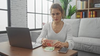 A focused young woman counts israeli shekels at home, seated by a laptop in a bright, cozy room. clipart