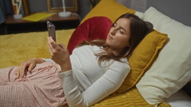 Genç, çekici bir kadın rahat bir yatak odasında uzanmış, akıllı telefonuna bakıyor, evde rahat bir ortam yaratıyor..
