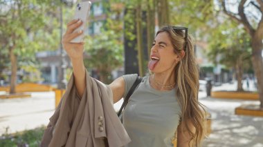 Neşeli genç bir kadın güneşli bir şehir parkında selfie dilini çıkarıyor, neşe ve günlük yaşam sergiliyor..