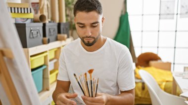 Sanat atölyesinde boya fırçalarını inceleyen yakışıklı bir adam, yaratıcılığı ve eğitim ortamındaki konsantrasyonu somutlaştırıyor..