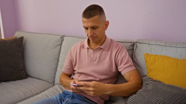 Oturma odasındaki kanepede oturan genç adam, cep telefonuyla meşgul, arka planda renkli yastıklar, rahat ve rahat bir iç mekan ortamı yaratıyor..