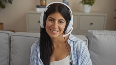 Esmer saçlı, güzel bir İspanyol kadın. Kulaklık takıyor. Oturma odasında oturuyor ve kameraya gülümsüyor..