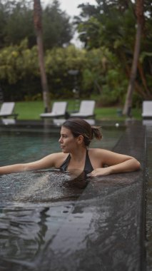 Dalgın genç bir kadın, bereketli yeşilliklerle çevrili huzurlu bir bali tatil köyünün sonsuz havuzunda dinleniyor..