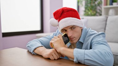 Yakışıklı, Noel Baba şapkalı, kendine güvenen bir görünüm ve güneş gözlüklü, rahat bir oturma odasında poz veren bir adam..