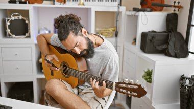 Sakallı genç bir adam müzik dolu bir stüdyo ortamında akustik gitar çalıyor..