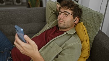 Sakallı, gözlüklü, kablosuz kulaklıklı, rahat, genç bir İspanyol adam akıllı telefonunu kullanarak evdeki bir kanepede dinleniyor..
