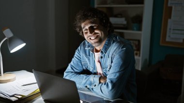 Yakışıklı İspanyol adam gece vakti iyi aydınlatılmış bir ofiste dizüstü bilgisayarın başında çalışırken gülümsüyor.
