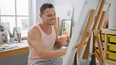 Parlak bir sanat stüdyosunda elinde kupayla tuvale bakan gülümseyen bir adam.