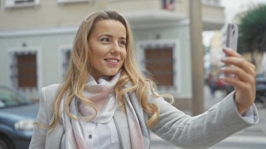 Gülümseyen genç bir kadın şehir caddesinde telefonuyla selfie çekiyor. Cazibe ve şehir cazibesi saçıyor..