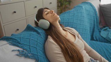 Uzun esmer saçlı, genç, çekici bir İspanyol kadın oturma odasında kulaklık takıyor ve mavi bir kanepede dinleniyor..
