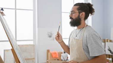 Önlüklü genç sakallı bir adam parlak bir sanat stüdyosunda boya fırçası tutuyor..