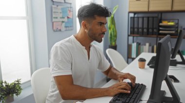 Modern ofis ortamında bilgisayarda çalışan genç İspanyol bir adam. Organize belgeler ve masanın üzerinde bir fincan kahve var..