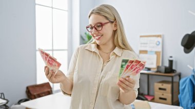 A young female with glasses smiles while examining israeli shekels in a bright office setting. clipart