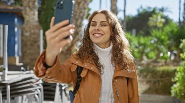 Parkta yeşillik içinde selfie çekerken gülümseyen genç İspanyol kadın.