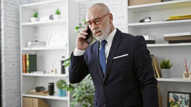 Takım elbiseli olgun işadamı modern ofis ortamında telefonla konuşuyor..