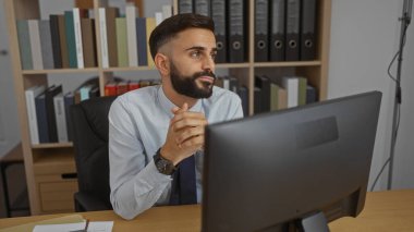 Ofis ortamında çalışan, bilgisayarlı bir masada oturan ve arka planda klasörleri olan yakışıklı İspanyol bir adam..