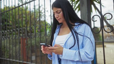 Güzel, genç, İspanyol bir kadın şehir dışında, çizgili bir gömlek giyiyor ve akıllı telefonuna bakıyor..