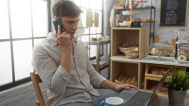 Fırında bir masada oturan genç adam, arka planda çeşitli fırınlanmış malların sergilendiği bir dizüstü bilgisayarda çalışırken telefonla konuşuyor.