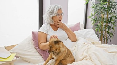 Yatağında köpeğiyle kır saçlı, bitkilerle dolu rahat bir yatak odasında şaşırmış yaşlı bir kadın.