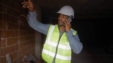 Yansıtıcı yelek giyen ve kasketli genç adam inşaat alanında telefonla konuşuyor. Görülmemiş bir şeye işaret ediyor. Liderlik ve iletişim duygusunu aktarıyor..