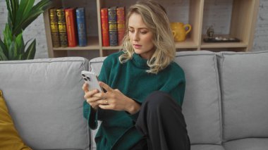 Beyaz, yeşil kazaklı genç bir kadın evdeki gri koltukta akıllı telefon kullanıyor.