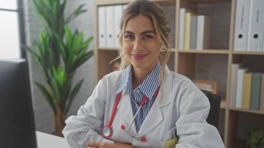 Modern bir klinikte steteskopla gülümseyen beyaz önlüklü, kendine güvenen sarışın bir kadın..