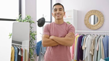 Kendine güvenen İspanyol bir adam, modern bir giyim odasında çeşitli kıyafetlerle gülümsüyor, gündelik bir yaşam tarzını yansıtıyor..