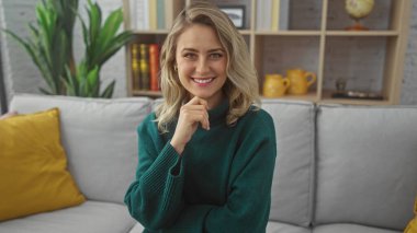 Gülümseyen, yeşil kazaklı, renkli yastıkları ve kitaplığı olan, modern bir oturma odasında oturan sarışın kadın.