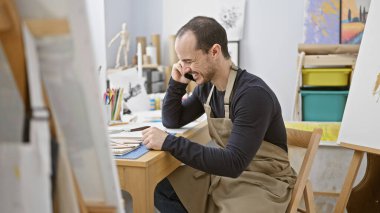 Bir sanat stüdyosunda önlük giymiş İspanyol kel bir adam telefonla konuşuyor.