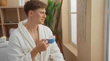 Genç adam spa merkezinde kahve içiyor, beyaz bir bornoz giyiyor, pencerenin dışına barışçıl bir şekilde bakıyor, sağlıklı bir atmosfer sergiliyor..