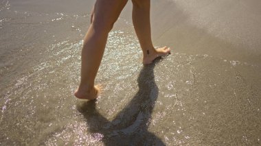 Güneşli bir sahilde, kumsalda çıplak ayakla yürüyen bir kadın. Sıcak bir günde güneş ışığında parıldayan bir su.